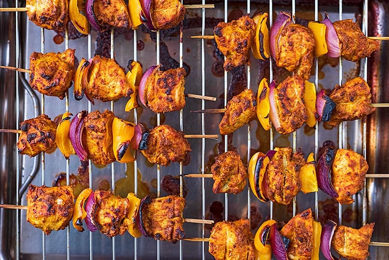 A cooking rack with eight cooked chicken tikka kebabs on it.