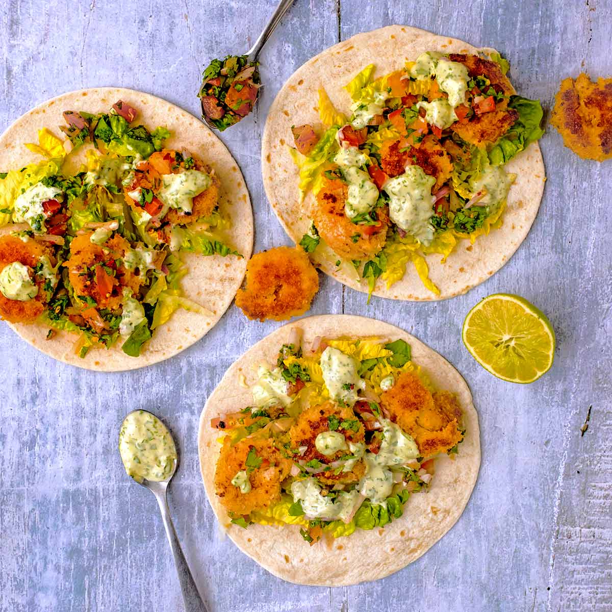 Three Crispy Prawn Tacos surrounded by lime wedges and spoons with salsa and sauce.