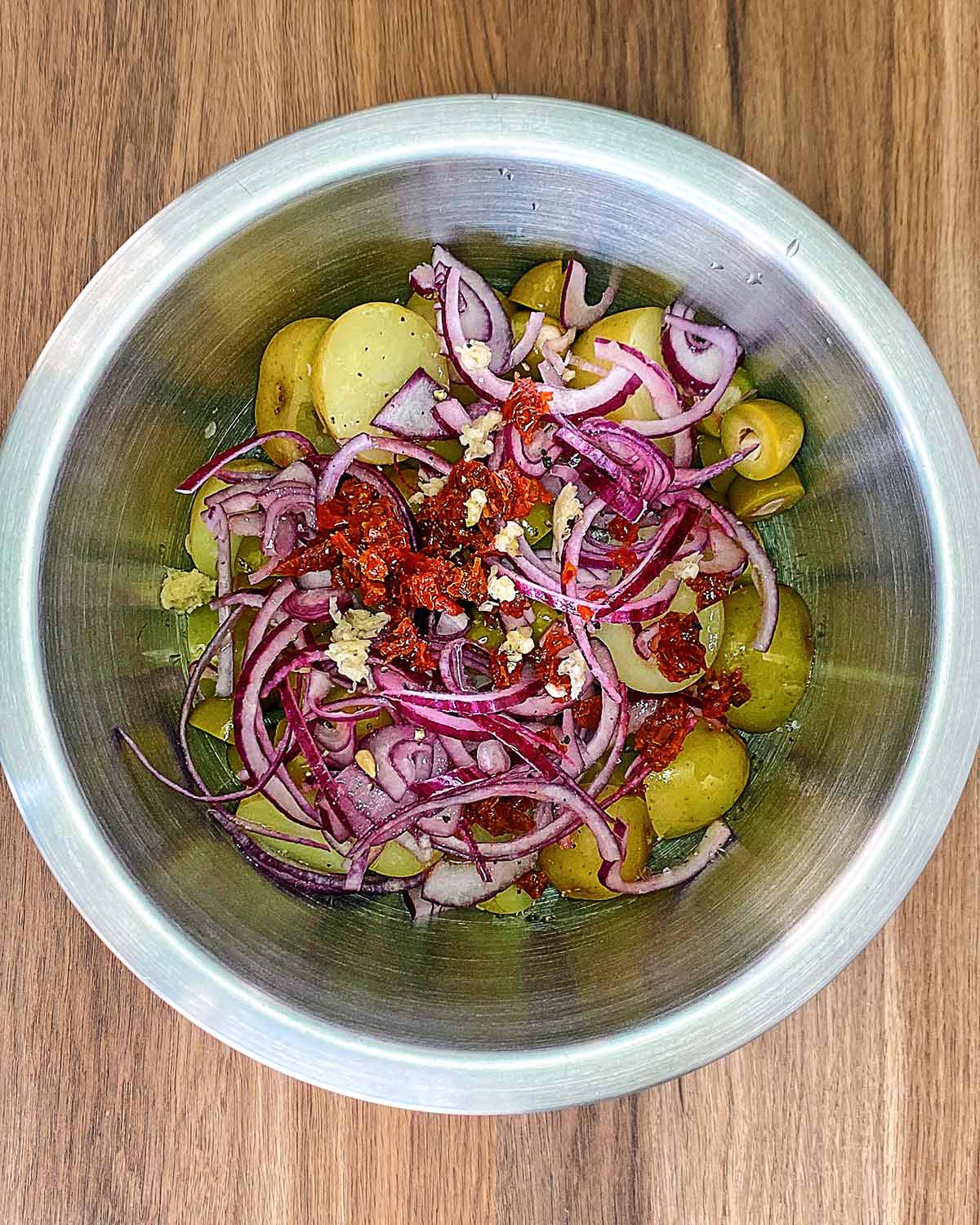 The cooked potatoes in a mixing bowl with onion slices, olives, tomatoes snd crushed garlic.