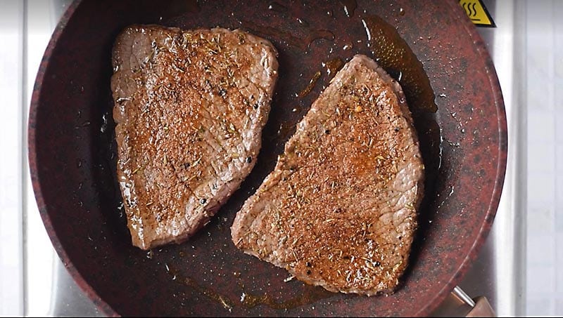 Two pieces of steak cooking in a frying pan.