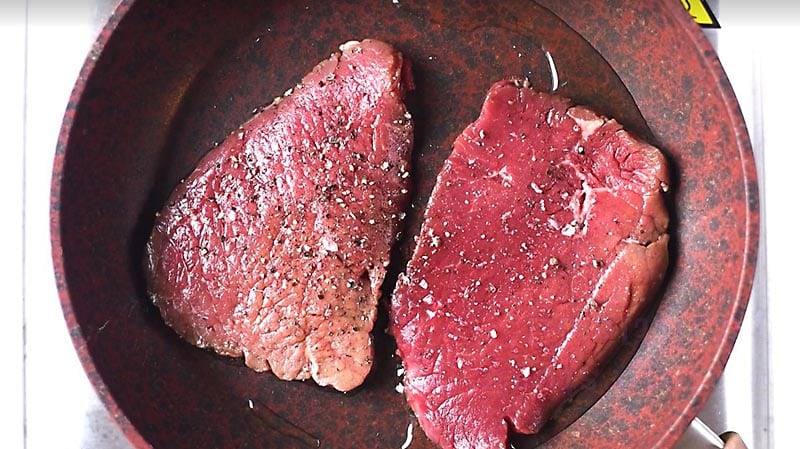 Two pieces of steak frying in a pan.
