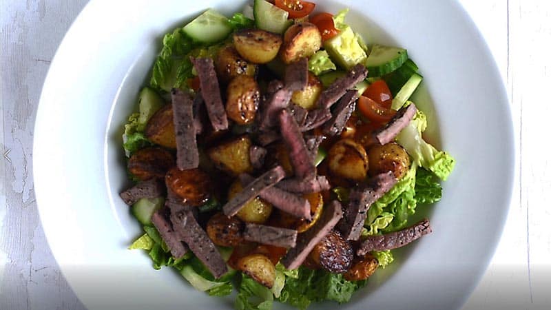 A large bowl containing lettuce, cucumber, tomato, potatoes and strips of steak.