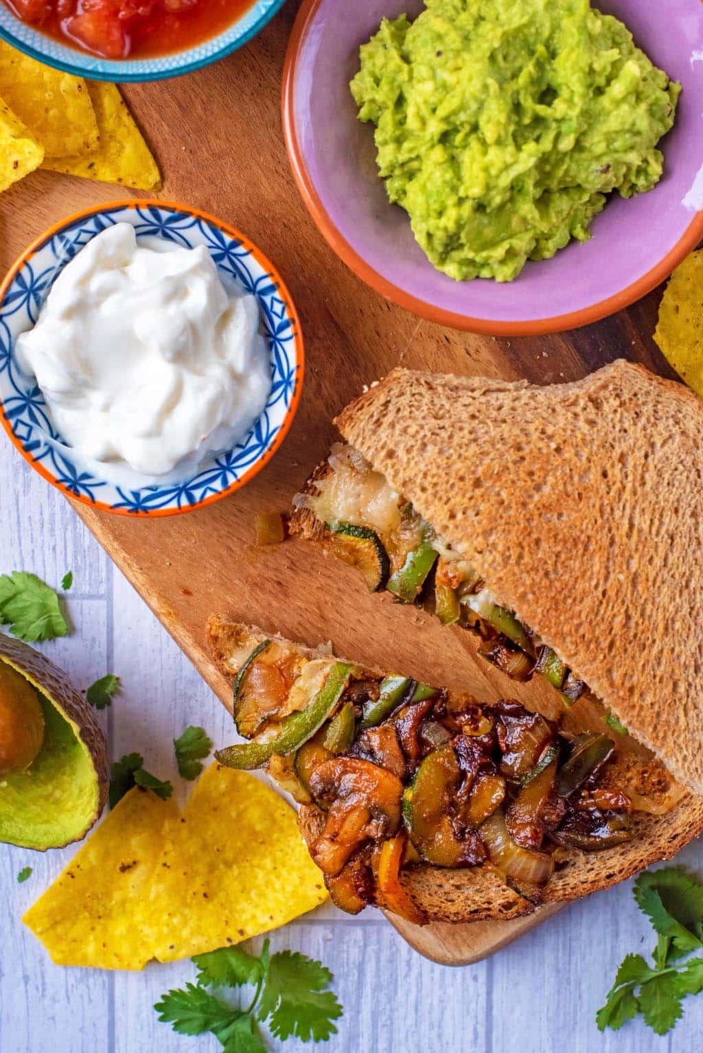 Toasted vegetable sandwich on a board with small bowls of guacamole and sour cream.