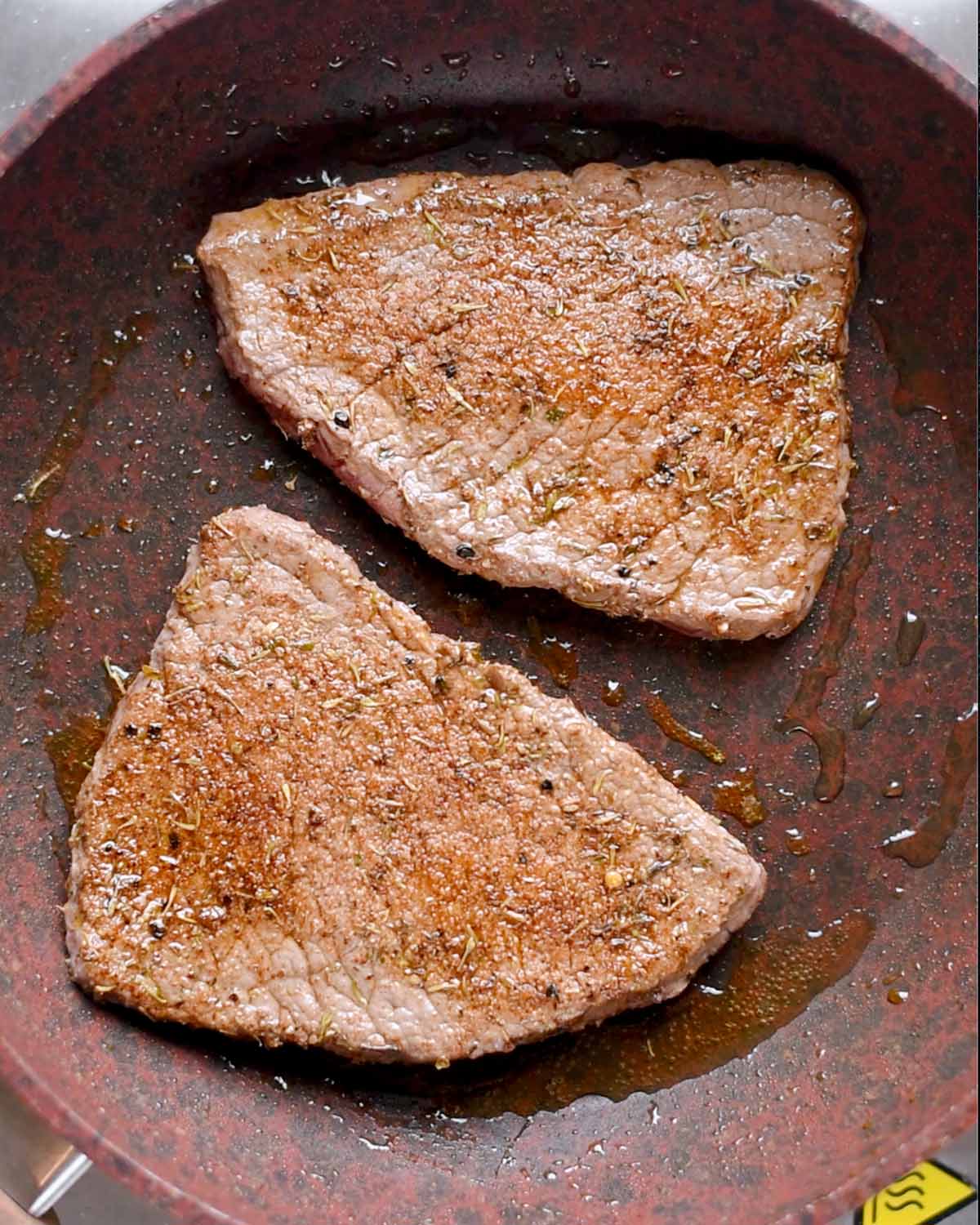 Two pieces of steak cooking in a frying pan.