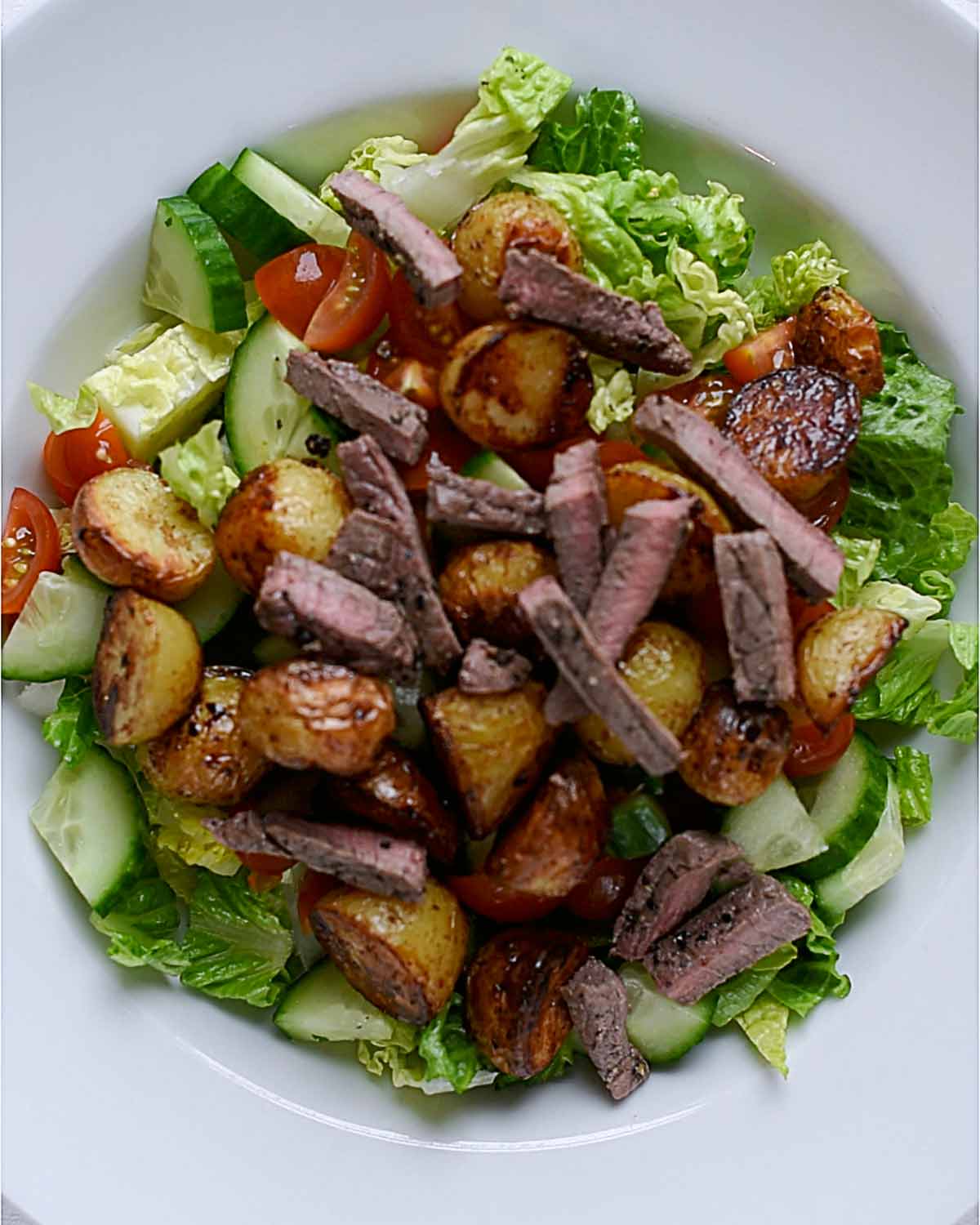 A large bowl containing lettuce, cucumber, tomato, potatoes and strips of steak.