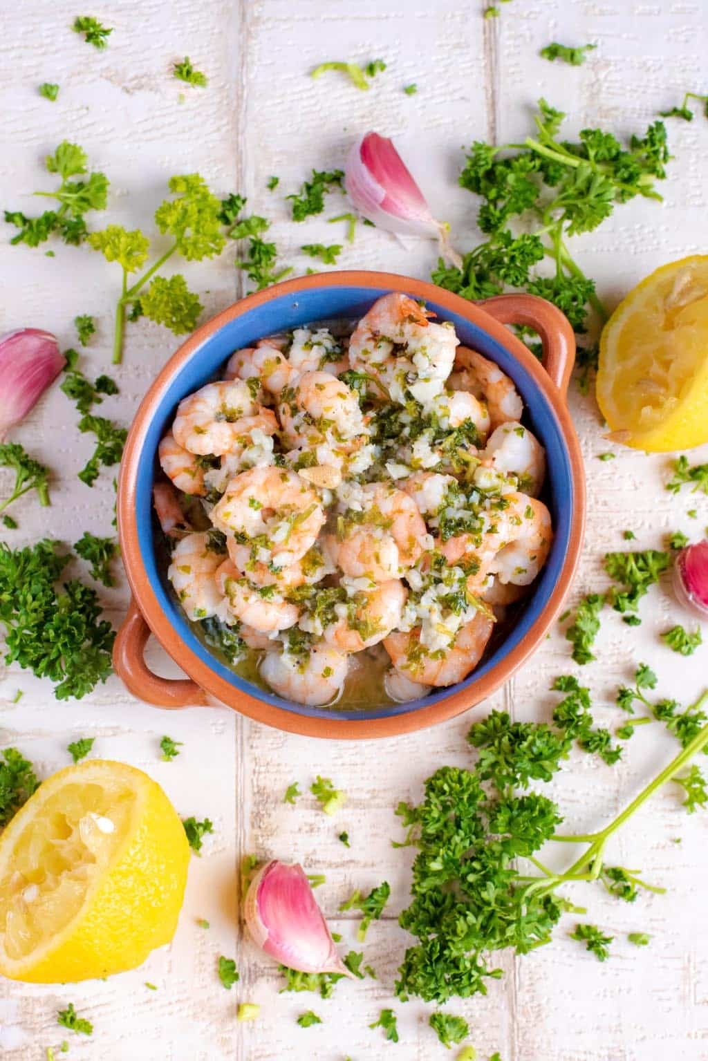 A bowl of cooked shrimp in garlic, herbs and oil, surrounded by parsley and lemon halves.