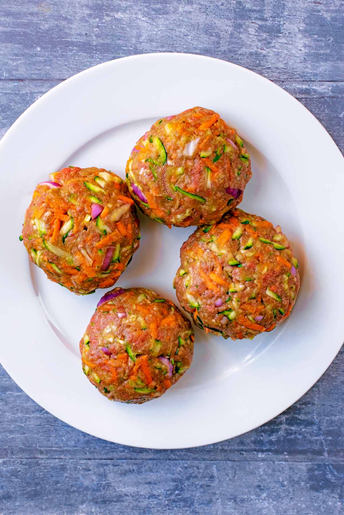 four uncooked beef and vegetable burger patties on a white plate.