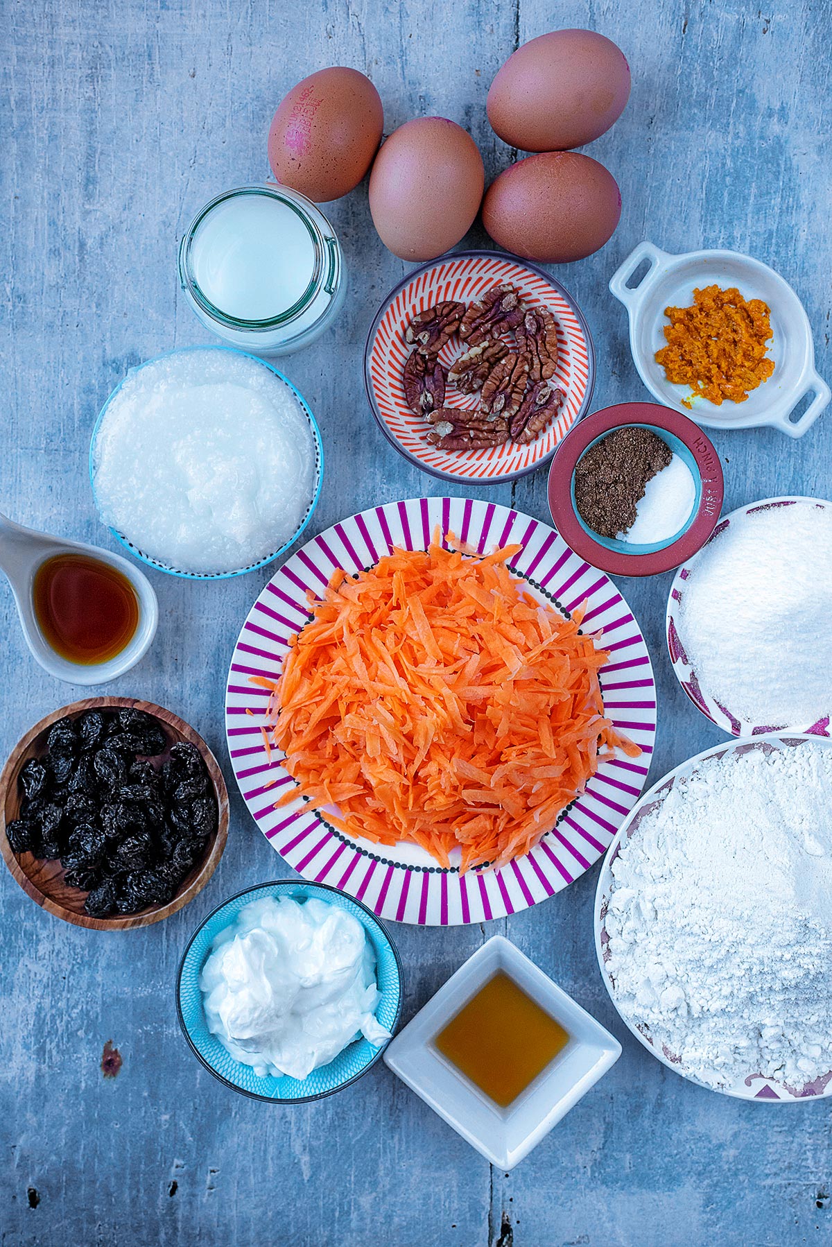 A plate of grated carrot surrounded by flour, sweetener, yogurt, raisins, nuts, spices, eggs, milk and orange zest