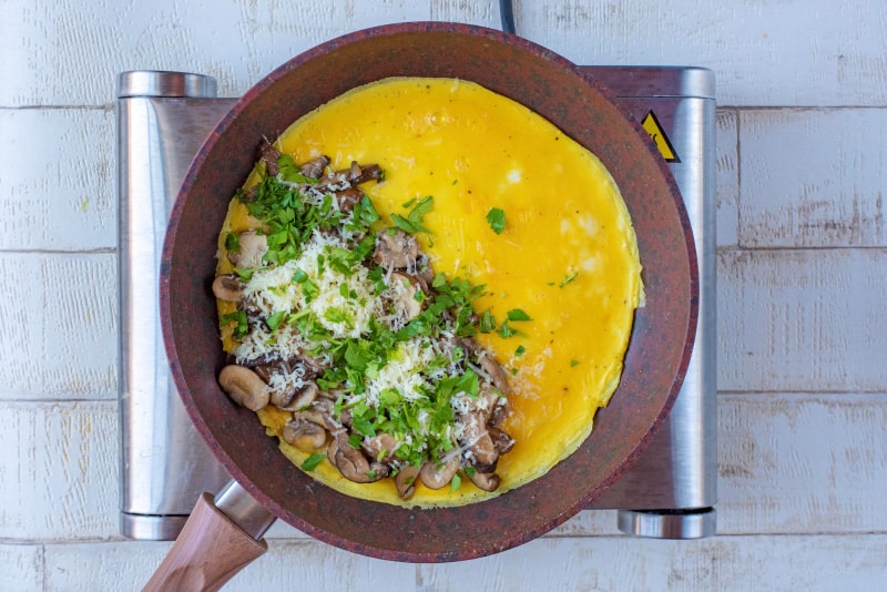A frying pan with a partially cooked omelette, half covered in mushrooms, greens and cheese.