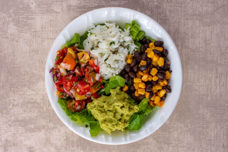 A white bowl containing shredded lettuce, rice, sweetcorn, black beans, salsa and guacamole.