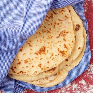 Easy Homemade Flatbreads wrapped in a blue gingham towel on a red surface. Flour is sprinkled around