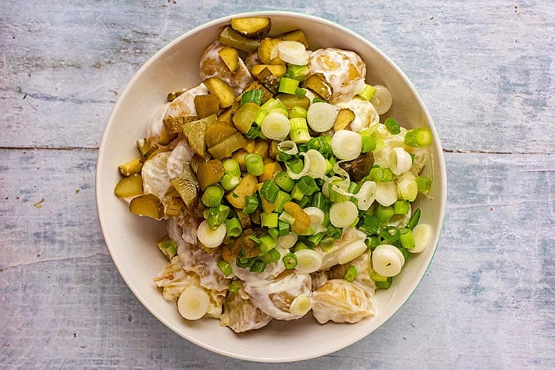 A bowl of potatoes covered in yogurt topped with scallions and gherkins.