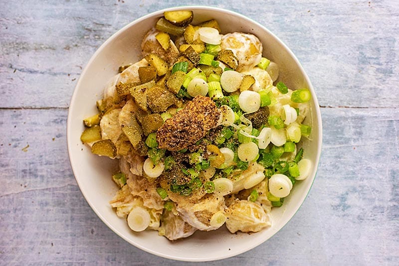A bowl of potatoes covered in yogurt topped with scallions, gherkins and mustard.