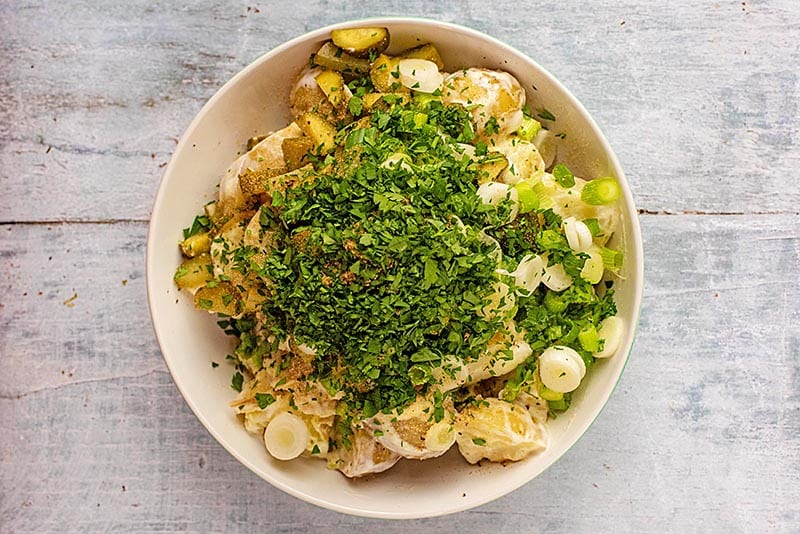 A bowl of potatoes covered in yogurt topped with scallions, gherkins, mustard and chopped parsley.