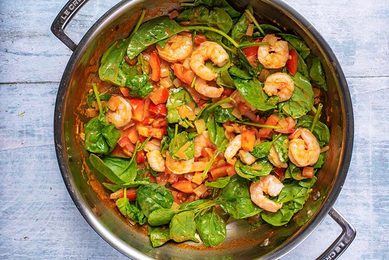 A large pan with prawns, tomatoes and spinach cooking in a curry sauce.