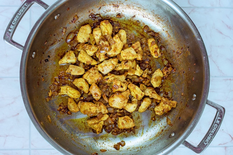 Diced chicken, chopped onions, garlic and spices cooking in a large silver pan.