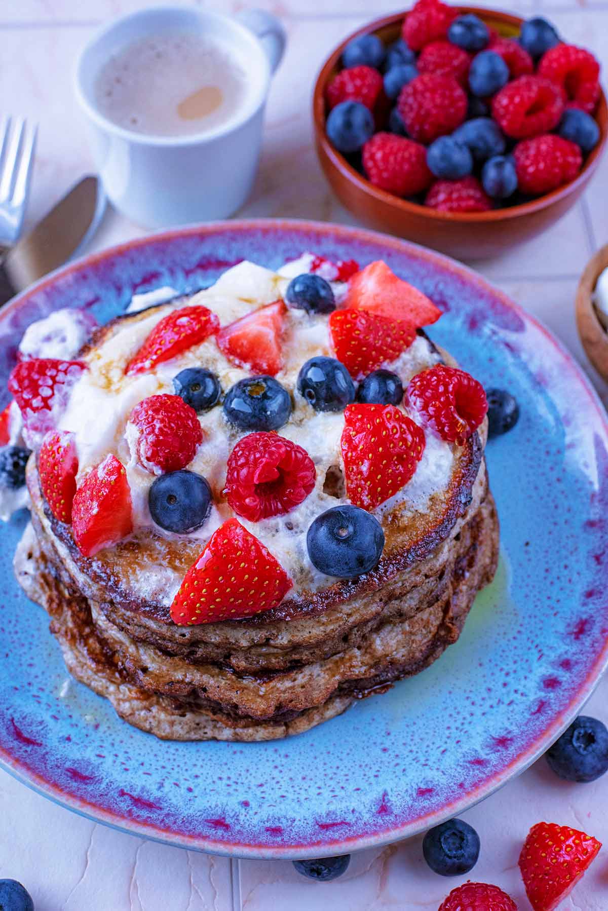 A plate of pancakes topped with cream and berries next to a bowl of more berries.