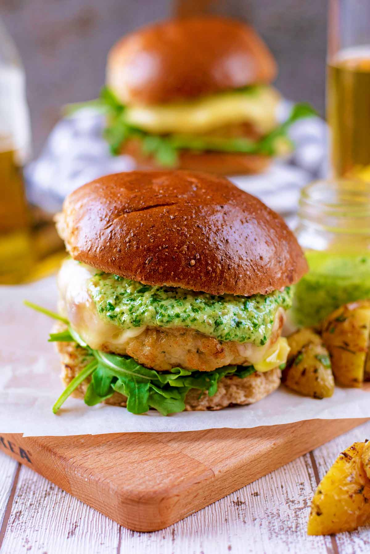 A chicken burger in a bun on a wooden board, beer and another burger in the background.