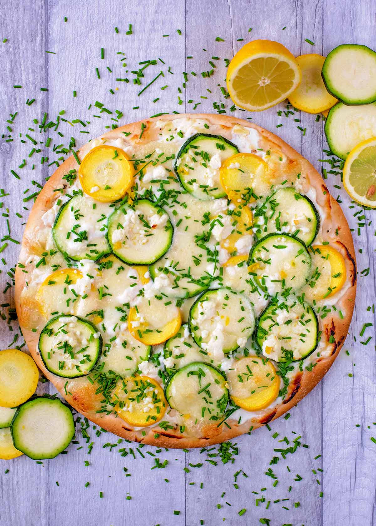 A courgette pizza on a wooden surface surrounded by chopped chives.