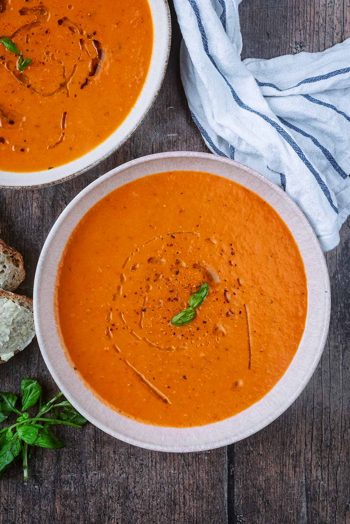 Bowls of red pepper and tomato soup next to a striped towel and bunch of basil leaves.