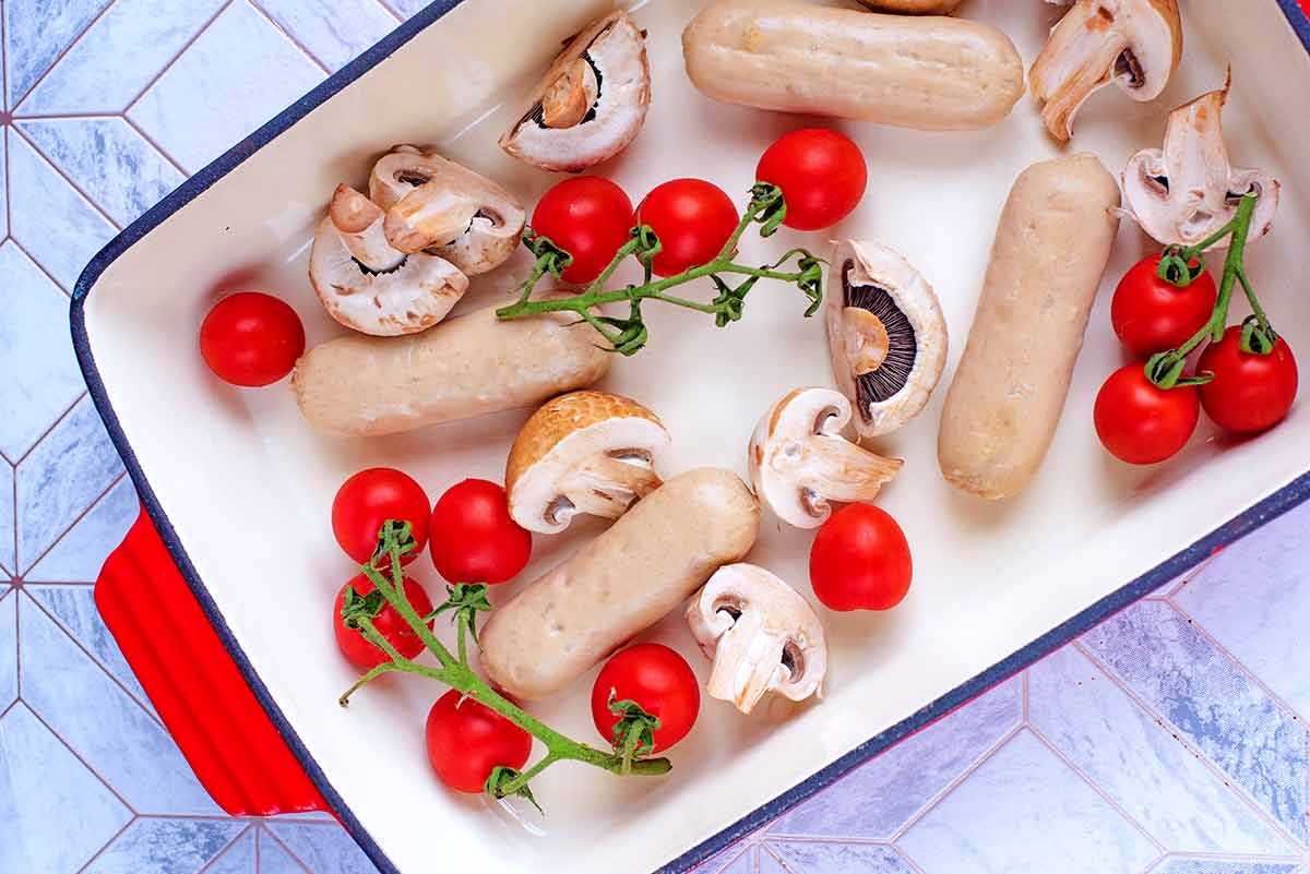 A large red baking dish containing sausages, halved mushrooms and cherry tomatoes.