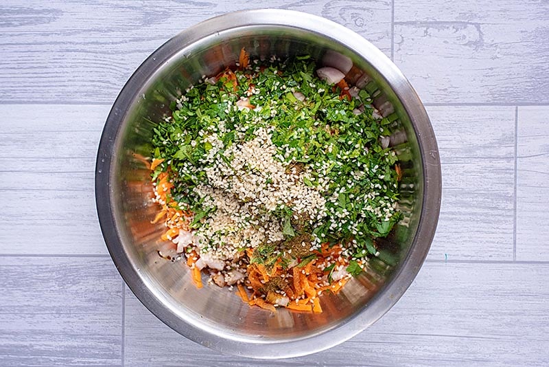 A large metal bowl full of grated carrot, chopped onion, cilantro and sesame seeds.