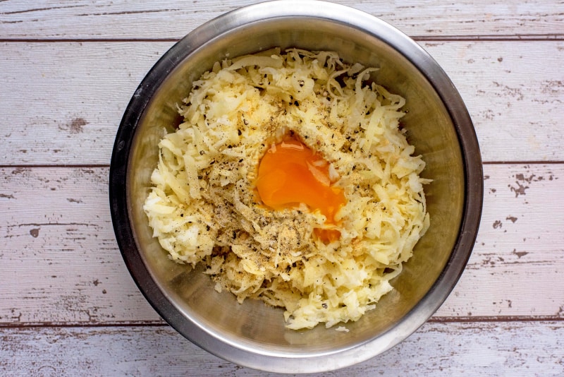 A stainless steel mixing bowl with grated potato, onion, an egg and seasoning.