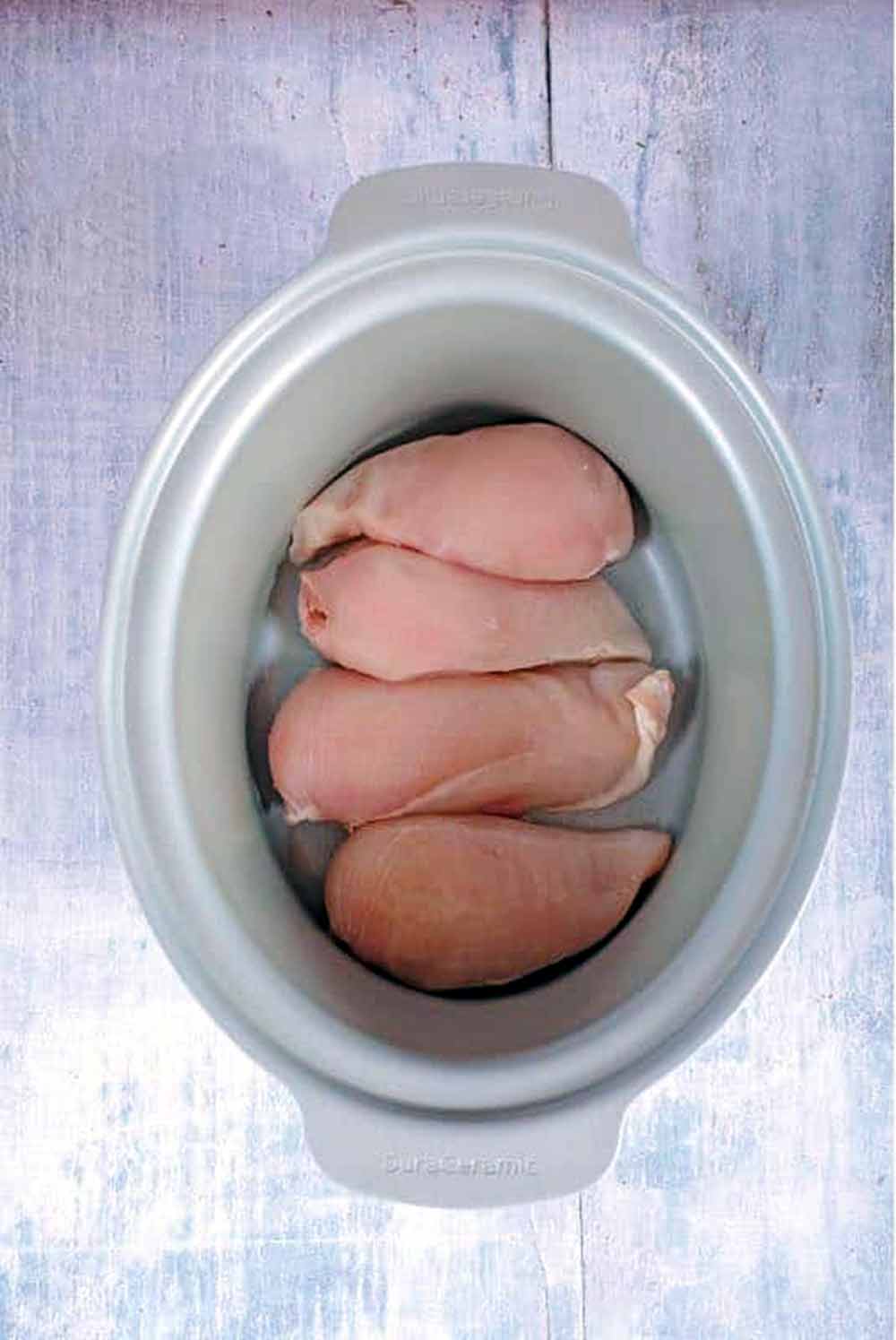 Four raw chicken breasts in a slow cooker pot.