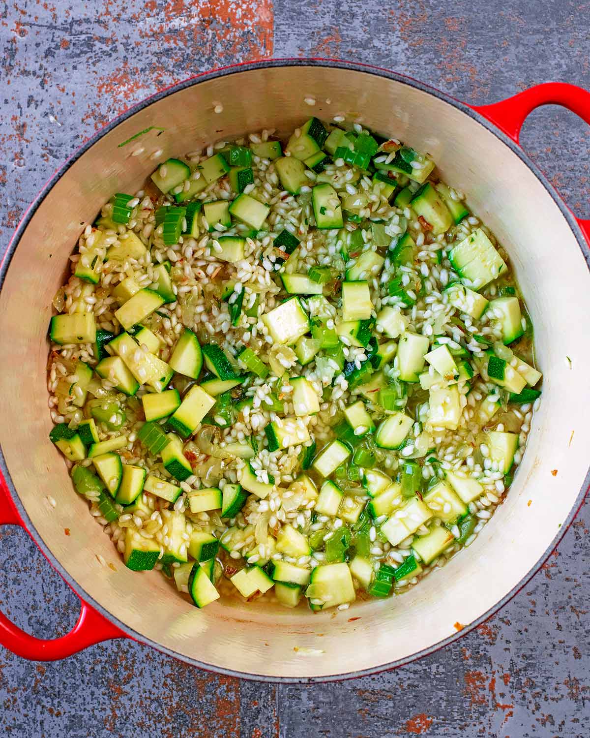 A large red pan with celery, shallots, courgette, risotto rice and stock.