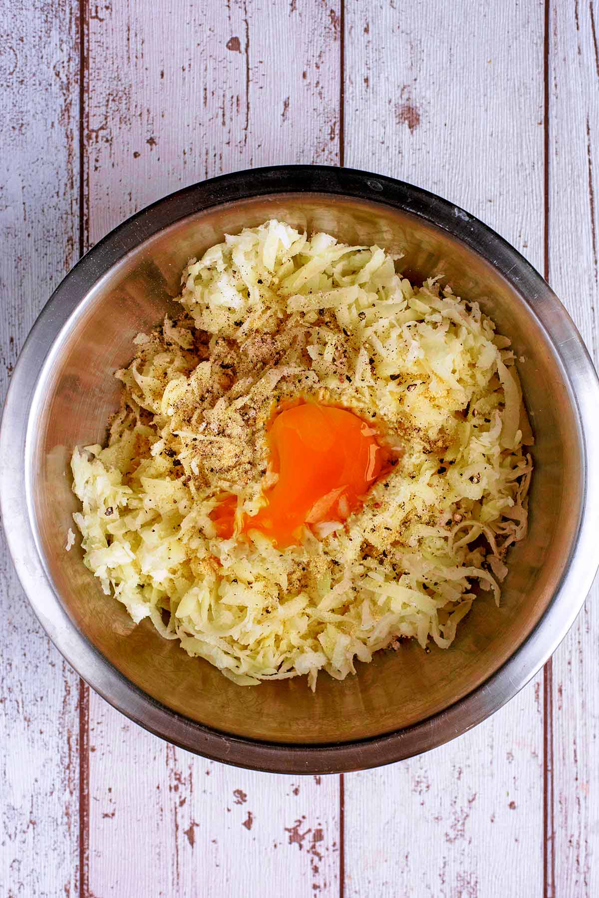 A stainless steel mixing bowl with grated potato, onion, an egg and seasoning.