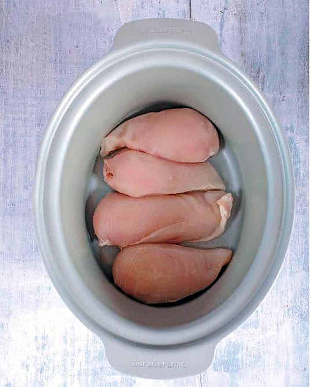 Four raw chicken breasts in a slow cooker pot.