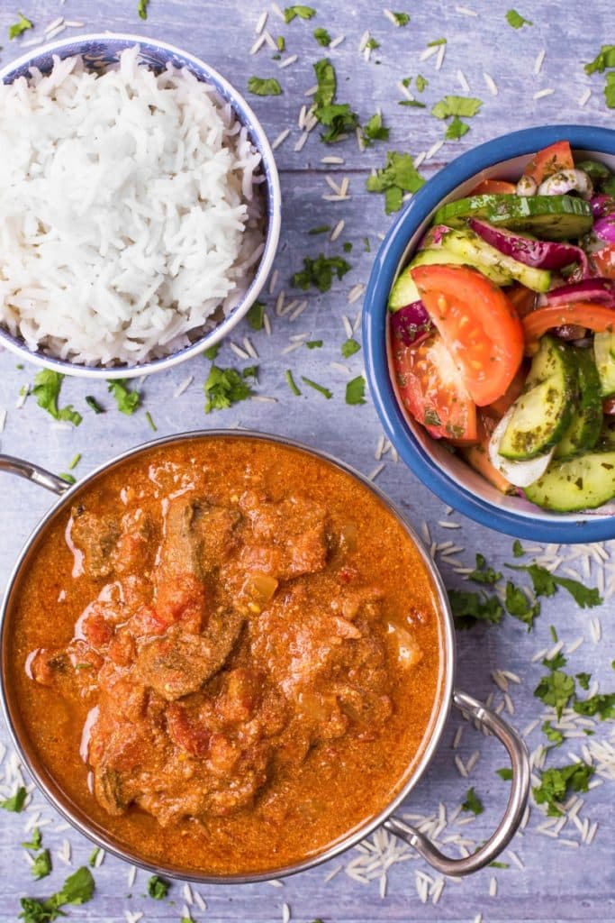 Beef curry in a balti dish next to a bowl of rice and a side salad