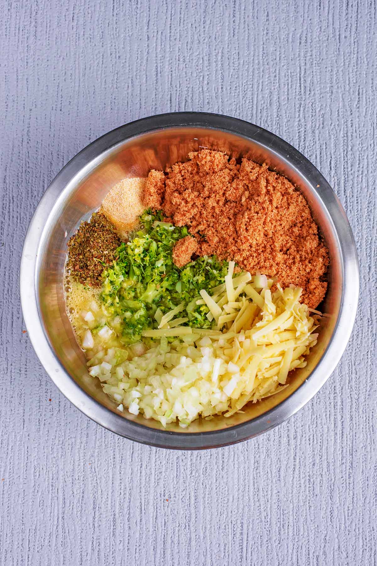 A mixing bowl containing mashed broccoli, breadcrumbs, grated cheese, chopped onion and herbs.