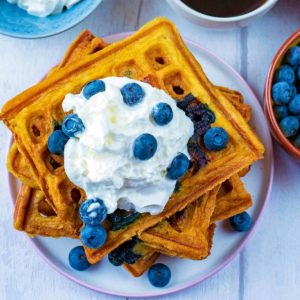 A pile of Blueberry Waffles on a white plate with cream and blueberries on top.
