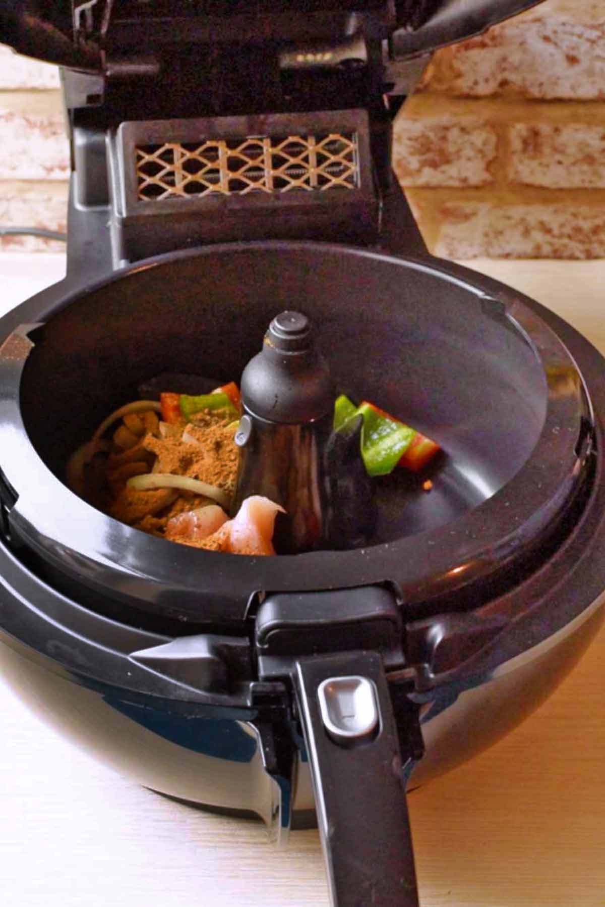 An air fryer with the lid open containing raw chicken, vegetables and spices.
