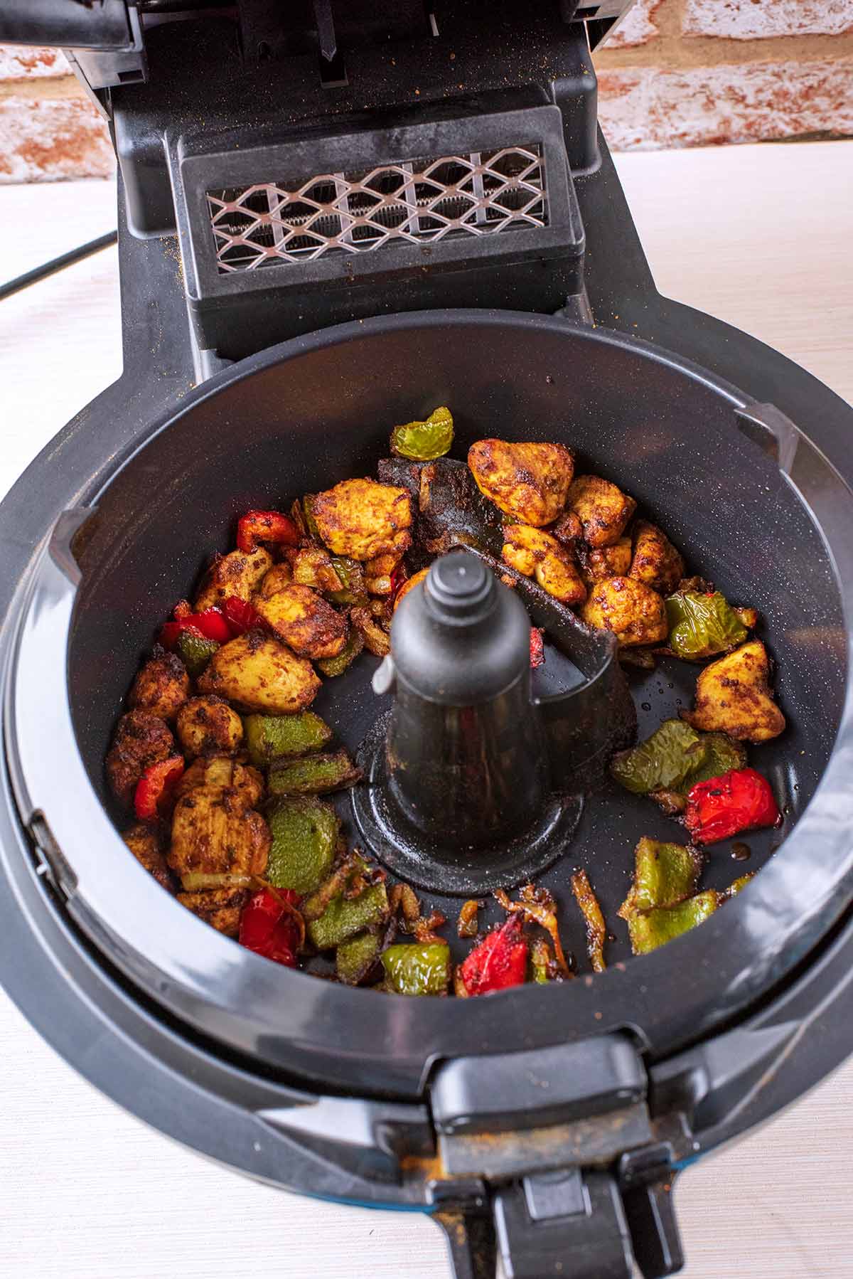 An air fryer with the lid open containing cooked chicken, vegetables and spices.