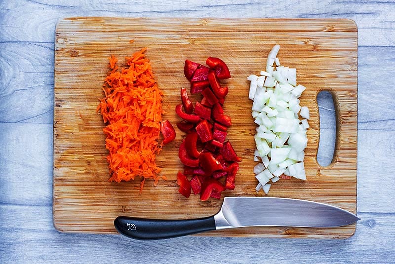 A wooden chopping board with grated carrot and diced red pepper and onion.