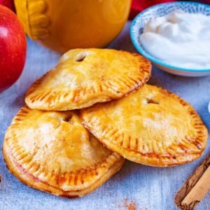 Three apple hand pies surrounded by an apple, cinnamon sticks, yogurt and a cup of coffee.