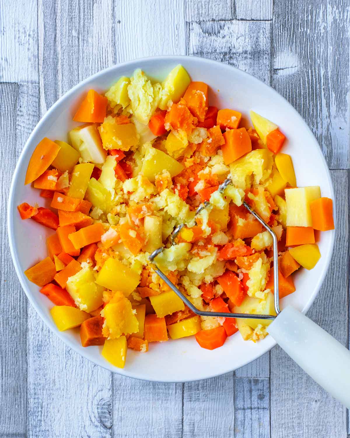 A selection of cooked root vegetables being mashed in a white bowl.