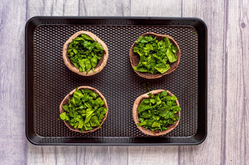 Four upturned portobello mushrooms topped with chopped spinach on a black baking tray.