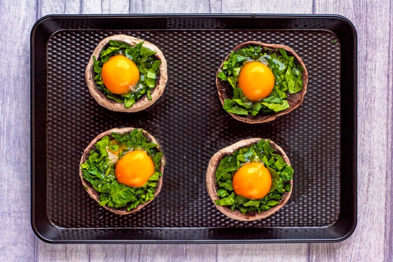 Four upturned portobello mushrooms topped with chopped spinach and a cracked egg on a black baking tray.