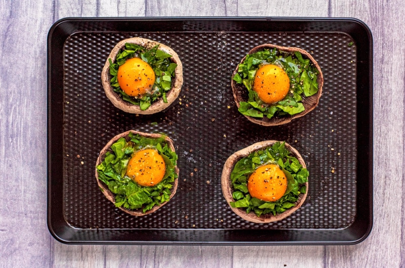 Four baked portobello mushrooms topped with chopped spinach and egg on a black baking tray.