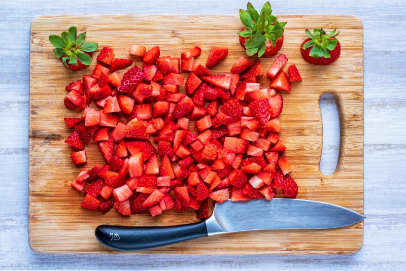 a wooden chopping board with chopped strawberries and a large knife.