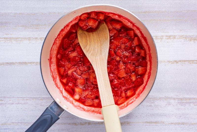 a wooden spoon mashing cooked strawberries in a pan.