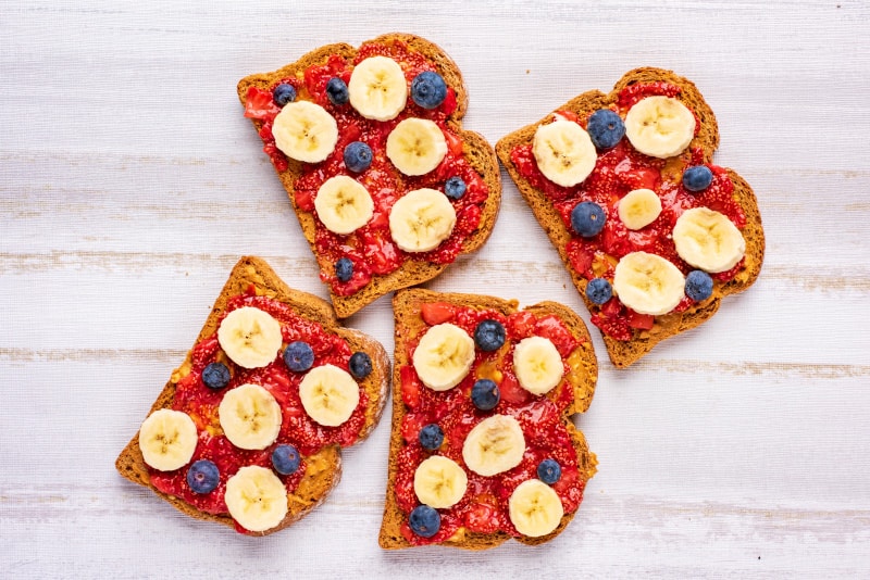 four slices of brown bread covered with peanut butter, strawberry jelly, banana slices and blueberries.