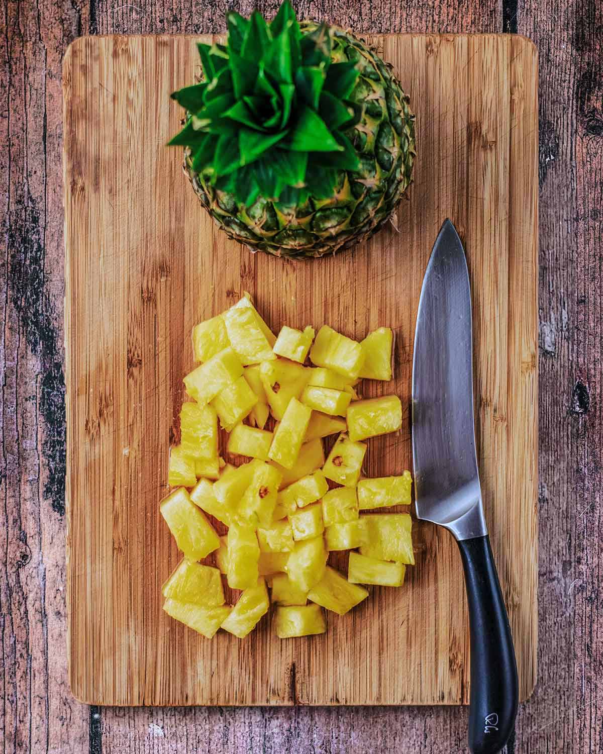 a wooden chopping board with a chef's knife, chunks of pineapple and a half whole pineapple.