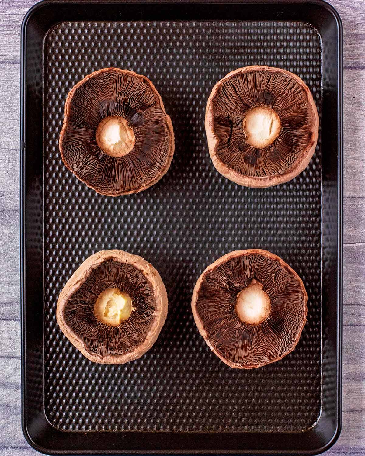 Four Portobello mushrooms on a black baking tray.