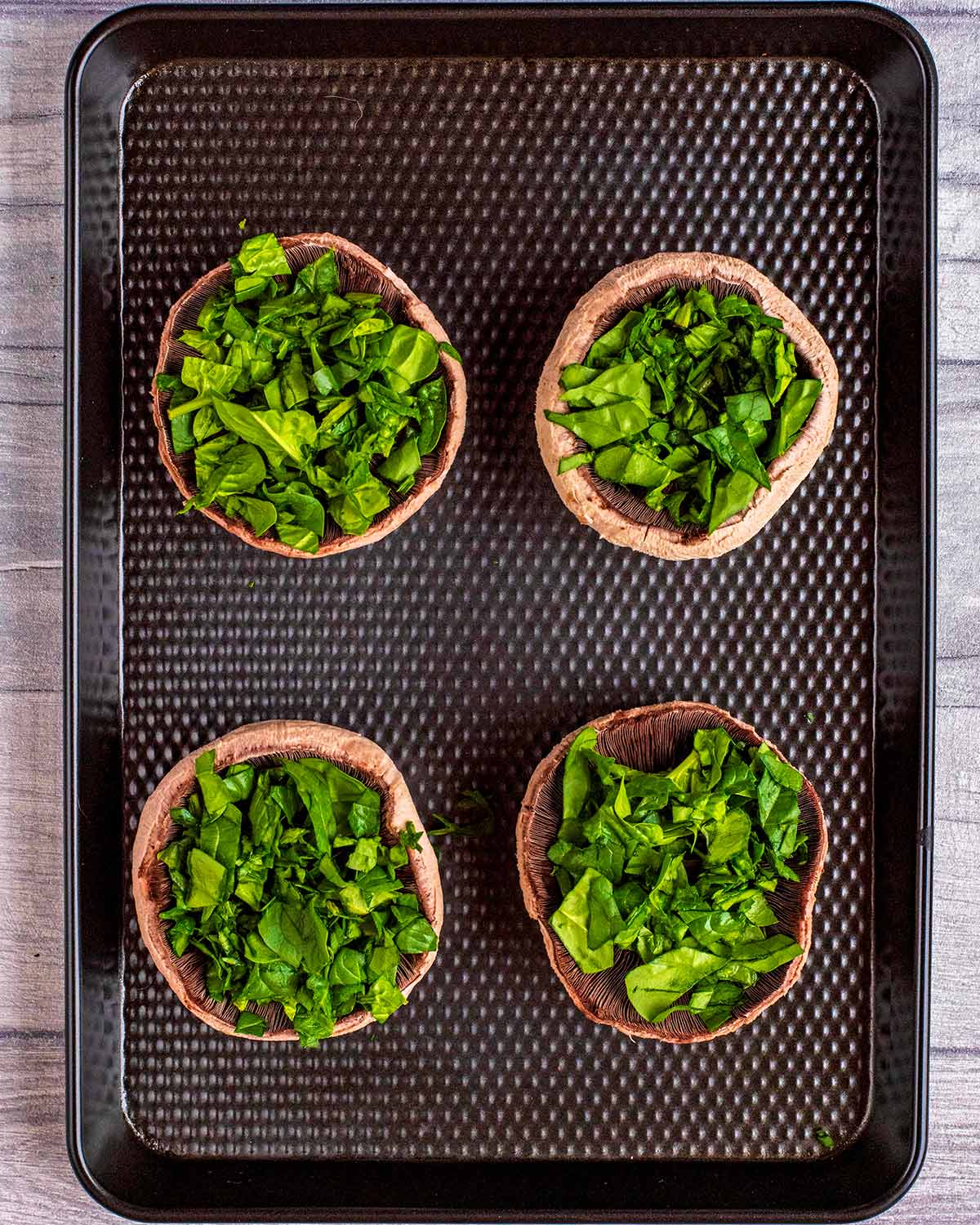 Four upturned portobello mushrooms topped with chopped spinach on a black baking tray.