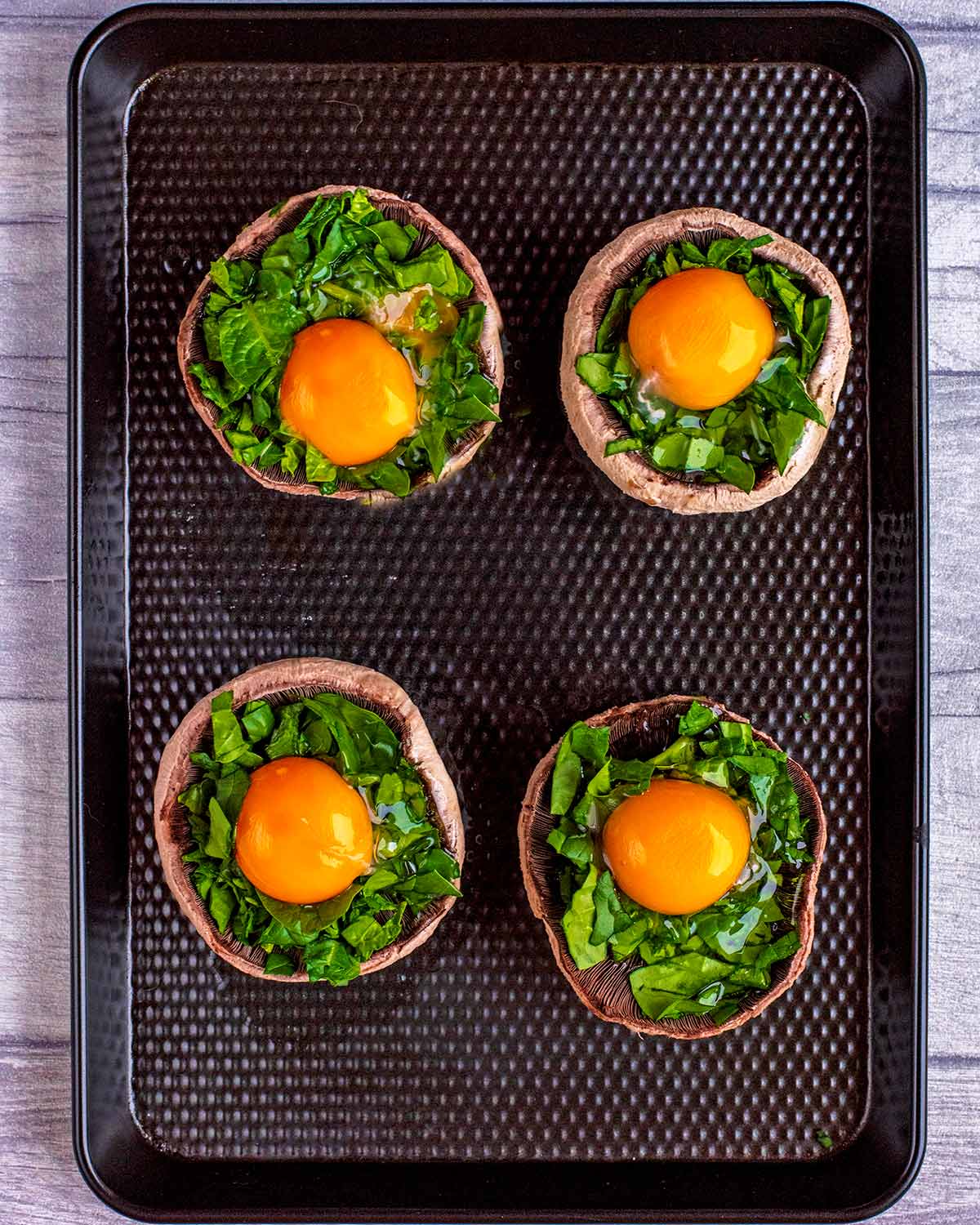 Four upturned portobello mushrooms topped with chopped spinach and a cracked egg on a black baking tray.