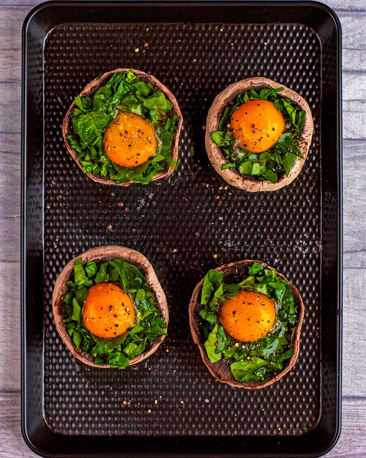 Four baked portobello mushrooms topped with chopped spinach and egg on a black baking tray.